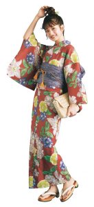 赤地にイエローやブルーなど、色とりどりな花柄が描かれた浴衣を着たモデルの写真。正面から撮影。手にはセカンドバッグ型のあみあみバッグ、草履は巻と天がキャラメル色で、鼻緒が白。髪の毛は頭上でお団子にされており、緑のかんざしをさしている。
