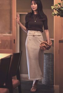 ブラウンのトップスに、淡いピンクのロングスカートを組み合わせ。ロングネックレスと、ベージュとオレンジが混ざったような色のバッグをセカンドバッグのように持っている。